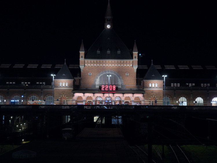 Vista nocturna de la estación de trenes. En la fachada hay un gran reloj digital
que marca las 22:08 y en una torre del edificio hay un reloj analógico más pequeño.
