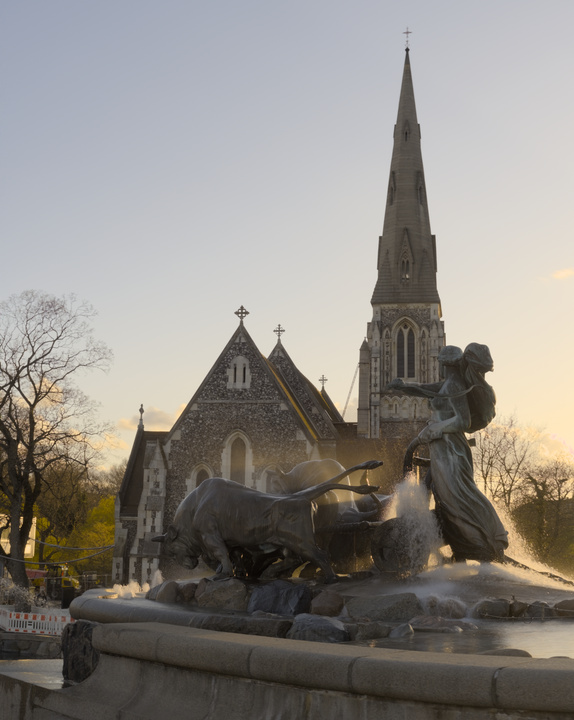 Fuente de agua con una estatua de una mujer en una carroza tirada por dos toros. De fondo se ve una iglesia y el cielo del atardecer.
