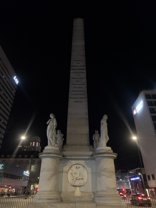 Foto nocturna de un obelisco rodeado de cuatro estatuas de estilo clásico grecorromano. En la base del obelisco hay un grabado con el mismo estilo de las estatuas.
