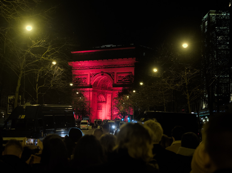 Arco del triunfo iluminado de rojo. En primer plano se ven una multitud de gente.
