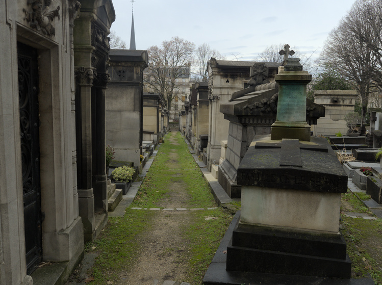 Pasillo de tierra de un cementerio rodeado por tumbas. El día está nublado y como es invierno, crece pasto en los costados del camino.
