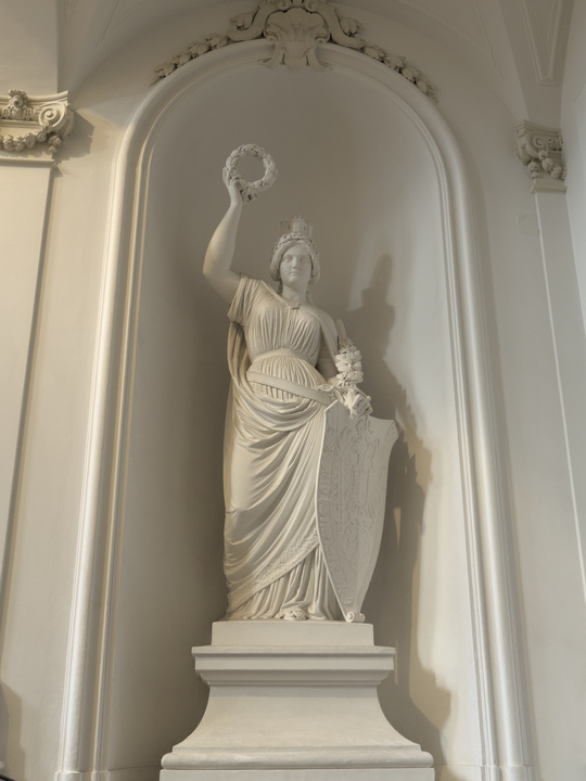 Estatua de una mujer con un escudo en una mano y una corona de laureles en alto en la otra.
